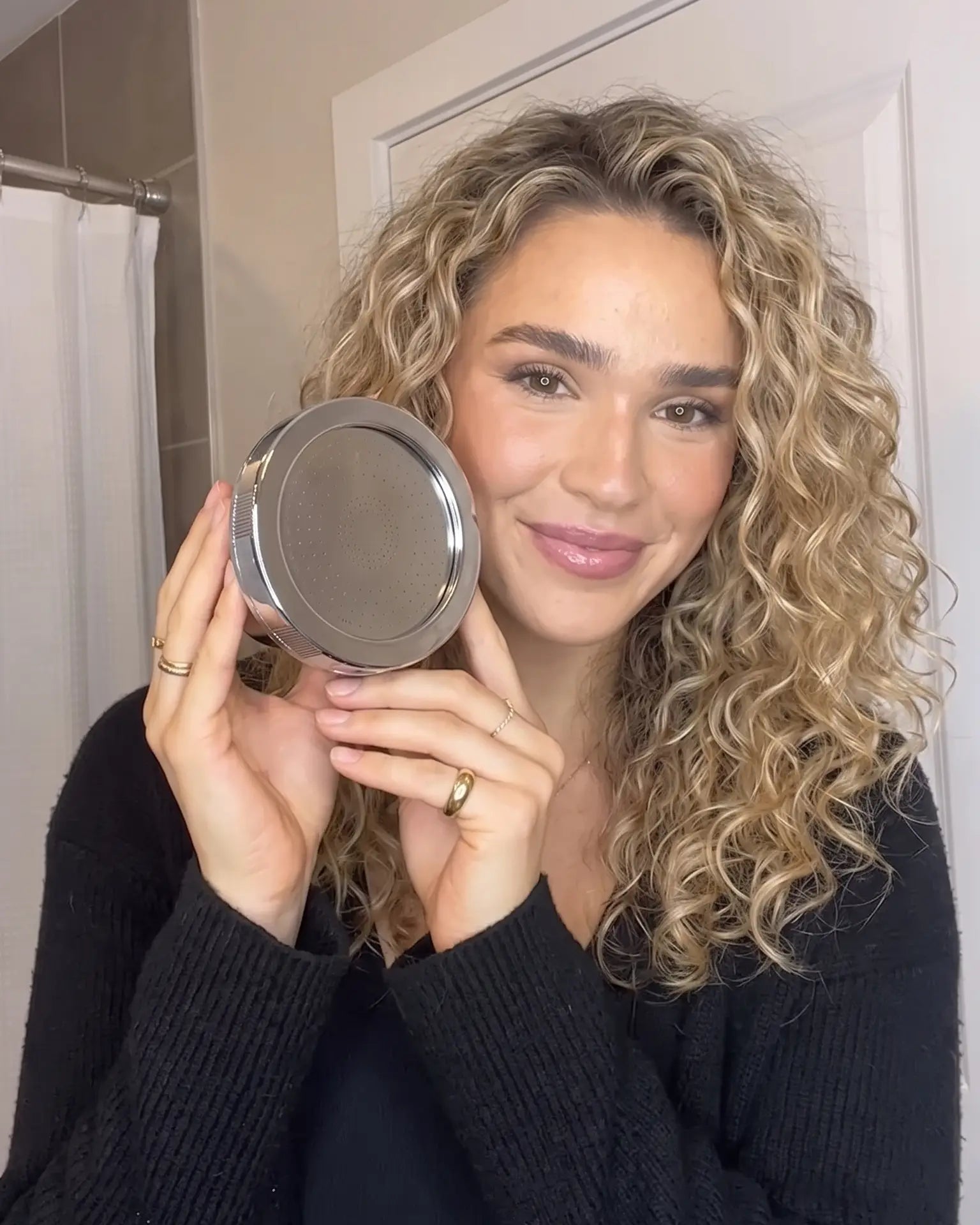 A woman with curly blonde hair holds up a round silver compact mirror in front of her face, wearing a black knit sweater in a bathroom setting.