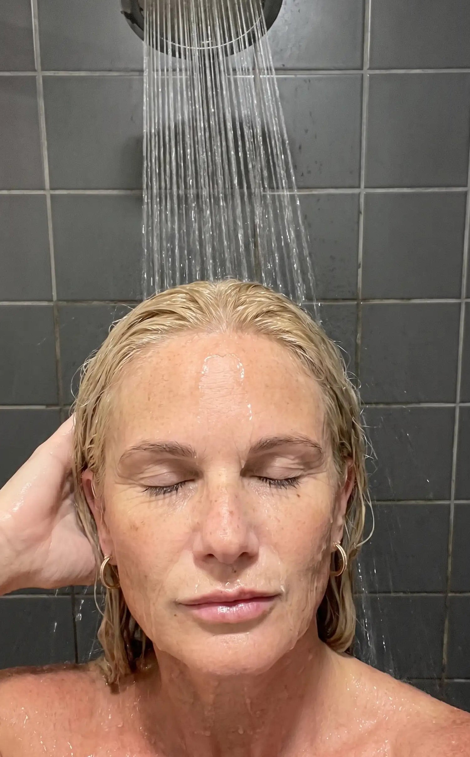 A woman with wet blonde hair stands in a shower under a rain headpiece, with water flowing over her face and shoulders against a dark tiled wall.